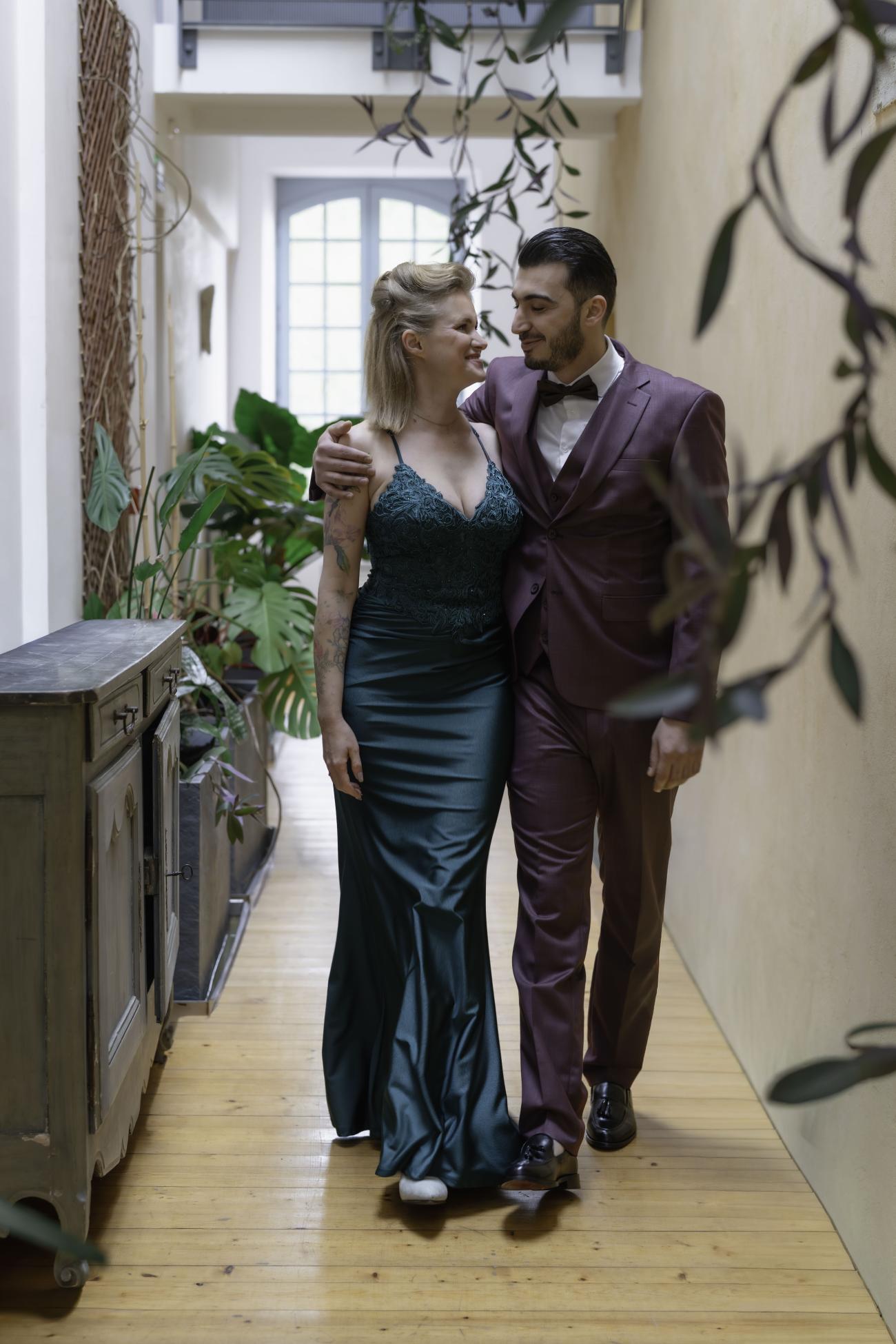 C'est un couple élégant, invité à un mariage haut de gamme au château Lacoste à Lupiac dans le Gers. Ils déambules dans un des couloirs du château, dans des vêtements très élégants. Elle en robe de cocktail en satin, d'unvert profond. Lui a un costume prune, une chemise blanche et un noeud de papillon noir. Ils se regardent amoureusement. 